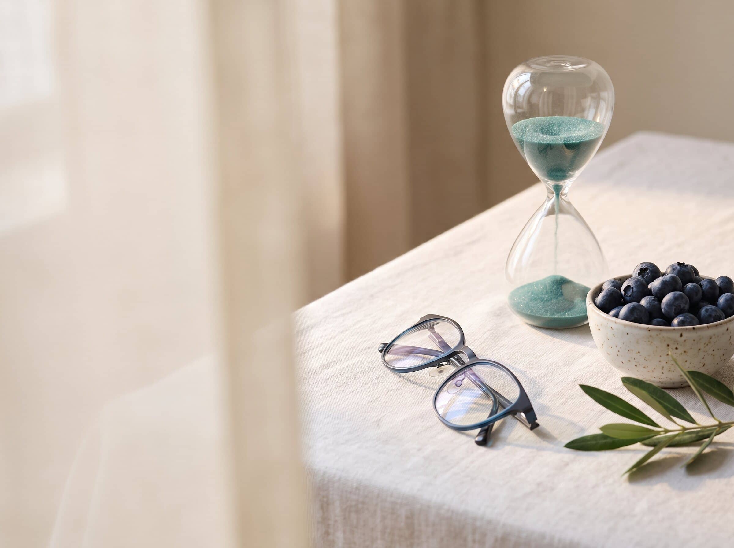 Still life of an hourglass with teal sand, blueberries, reading glasses and an olive branch on linen